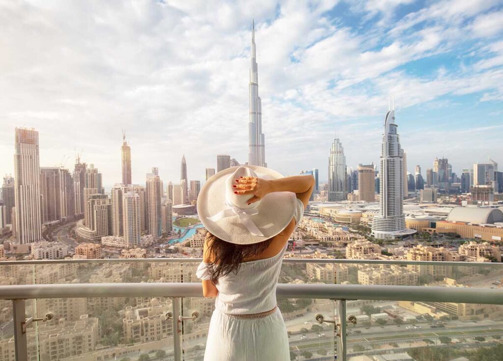 Woman with a white hat is standing on a balcony in front of dubai skyline